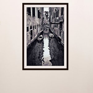 May include: Black and white framed print of a Venetian canal scene. The image shows a narrow waterway flanked by buildings, with boats and a bridge visible. The water reflects the architecture, creating a symmetrical composition.