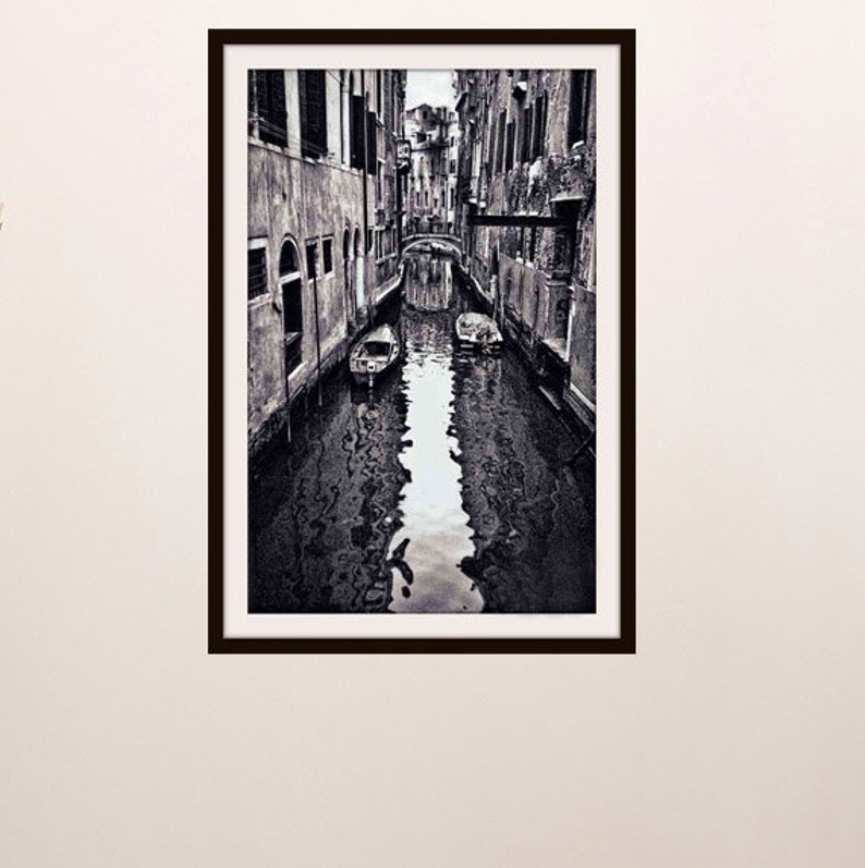 May include: Black and white framed print of a Venetian canal scene. The image shows a narrow waterway flanked by buildings, with boats and a bridge visible. The water reflects the architecture, creating a symmetrical composition.