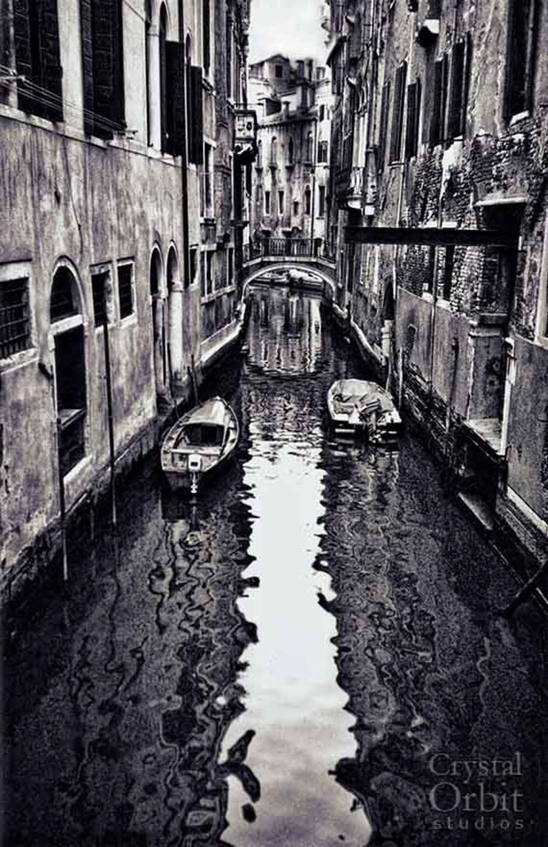 May include: Black and white photograph of a canal in Venice, Italy. The image shows narrow waterways flanked by aged buildings. Two small boats are moored in the water. A bridge spans the canal in the distance.