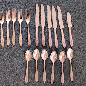 May include: A set of vintage silverware, including forks, knives, and spoons, arranged on a dark textured surface. The silverware has a copper-colored finish and a decorative pattern on the handles. The set includes six forks, six knives, and six spoons.