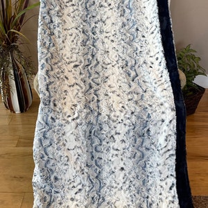 May include: A soft, white and blue faux fur throw blanket with a black border. The blanket has a wavy, abstract pattern.