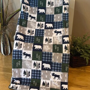 May include: A soft, plush fleece blanket with a patchwork design featuring bears, moose, and pine trees in shades of blue, green, and white.
