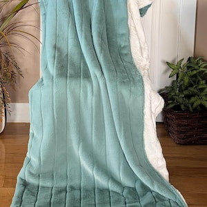 May include: A plush, dual-sided throw blanket featuring a soft, teal-colored faux fur top with vertical and horizontal channel stitching, contrasted by a white, textured, fleece-like underside that is visible along the folded edges.