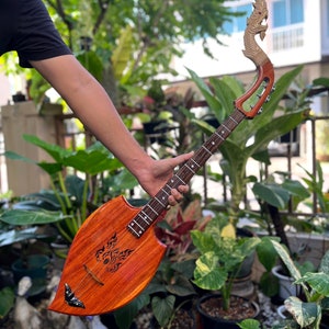 Puede incluir: Un instrumento musical tradicional tailandés llamado "Khim" con una cabeza de dragón de madera tallada y un cuerpo con forma de hoja. El instrumento está hecho de madera y tiene un acabado naranja natural. El cuerpo del instrumento está decorado con un diseño tallado de una criatura mítica.