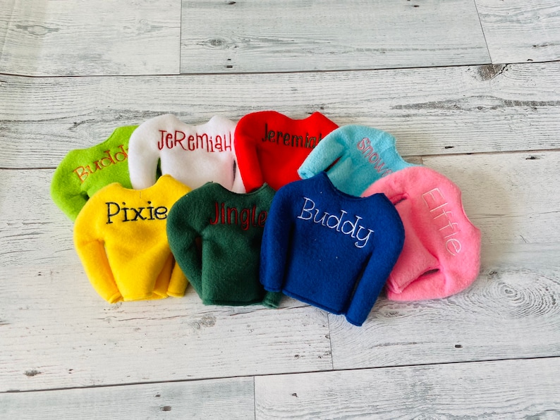 May include: Eight elf-sized sweaters in various colors, including green, yellow, blue, pink, red, white, and light blue. Each sweater has a different name embroidered on it, including Buddy, Pixie, Jingle, Buddy, Elfie, Jeremiah, Jeremiah, and Snow.