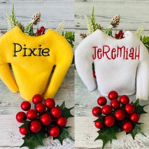 May include: Two elf-sized sweaters with personalized names. The yellow sweater says "Pixie" and the white sweater says "Jeremiah". Both sweaters are displayed on a wooden surface with red berries and green leaves.