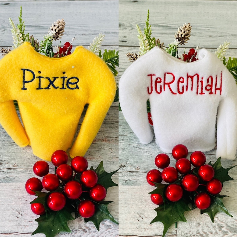 May include: Two elf-sized sweaters with personalized names. The yellow sweater says "Pixie" and the white sweater says "Jeremiah". Both sweaters are displayed on a wooden surface with red berries and green leaves.