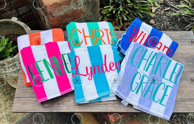 May include: A collection of personalized beach towels with vertical stripes in various colors, including orange, pink, green, blue, and purple. Each towel features a name embroidered in a contrasting color, such as "KENNEDE" and "WILLIAM".