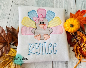 May include: A white embroidered shirt with a colorful turkey design and the name "Rylie" embroidered in blue. The turkey has a pink bow and a gray body. The shirt is surrounded by fall leaves and a small orange pumpkin.