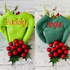 May include: Two green elf-sized sweaters with red embroidered text. One sweater is lime green with the word "Buddy" embroidered on it. The other sweater is forest green with the word "Jingle" embroidered on it.