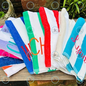 May include: A collection of colorful beach towels with striped patterns. Some towels feature embroidered names or initials in various colors, including pink, orange, and teal. The towels are folded and arranged on a wooden surface.