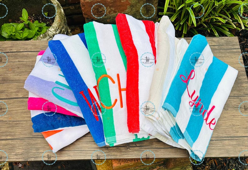 May include: A collection of colorful beach towels with striped patterns. Some towels feature embroidered names or initials in various colors, including pink, orange, and teal. The towels are folded and arranged on a wooden surface.