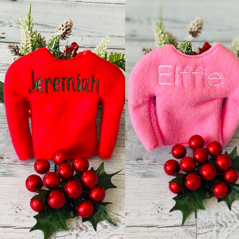May include: Two elf-sized Christmas sweaters, one red with the name "Jeremiah" embroidered in green, and one pink with the name "Elfie" embroidered in white. Both sweaters are on a white wooden background with a sprig of holly with red berries.