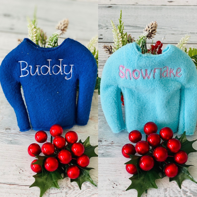 May include: Two elf-sized sweaters, one blue with the word "Buddy" embroidered in white, and one light blue with the word "Snowflake" embroidered in pink. Both sweaters are on a white wooden surface with a sprig of red berries and green leaves.