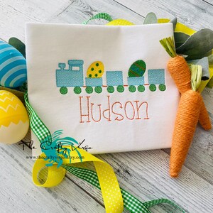 May include: A white t-shirt with an embroidered Easter train design featuring a blue engine, green boxcars, and yellow and green Easter eggs. The name "Hudson" is embroidered below the train.