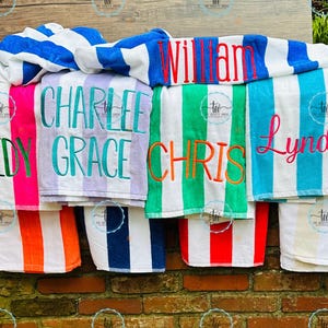 May include: A collection of personalized beach towels with vertical stripes in various colors, including pink, green, blue, and orange. Each towel is embroidered with a name in a contrasting color. The towels are displayed on a brick wall.