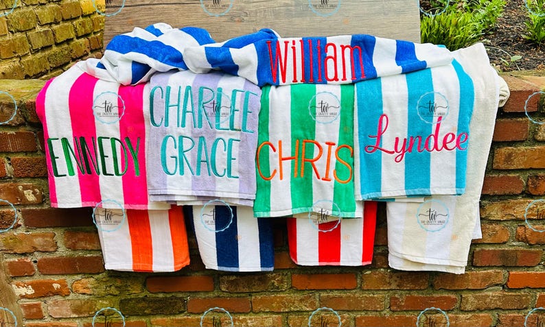 May include: A collection of personalized beach towels with vertical stripes in various colors, including pink, green, blue, and orange. Each towel is embroidered with a name in a contrasting color. The towels are displayed on a brick wall.