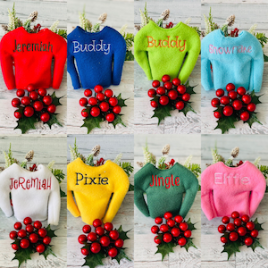 May include: Eight elf-sized Christmas sweaters in various colors with personalized names: Jeremiah, Buddy, Snowflake, Jeremiah, Pixie, Jingle, Elfie. Each sweater is displayed on a white wooden surface with a sprig of holly and red berries.