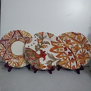 May include: Three round ceramic plates displayed on dark brown stands. The left plate features a geometric star pattern in orange, red, and cream. The center plate shows a botanical motif with pomegranates. The right plate has a leafy vine design.