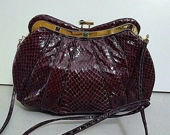 May include: A dark burgundy handbag with a textured snakeskin-like pattern, featuring a pleated body and a gold-tone metal frame closure. The bag includes a thin matching shoulder strap and a small green accent detail on the central clasp.