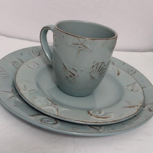 May include: A light blue ceramic mug and two plates with seashell designs. The mug sits on a smaller plate, which is on top of a larger plate. The plates and mug have a distressed, rustic look.
