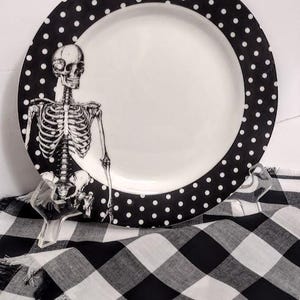 May include: A round ceramic plate featuring a white center and a wide black rim decorated with white polka dots. A detailed black and white illustration of a human skeleton is positioned on the left side of the plate, extending from the rim.
