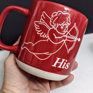 May include: Red ceramic mug with a white base and handle. The mug features a white illustration of Cupid with a bow and arrow, and the word "His" in white. The mug is held in a hand.