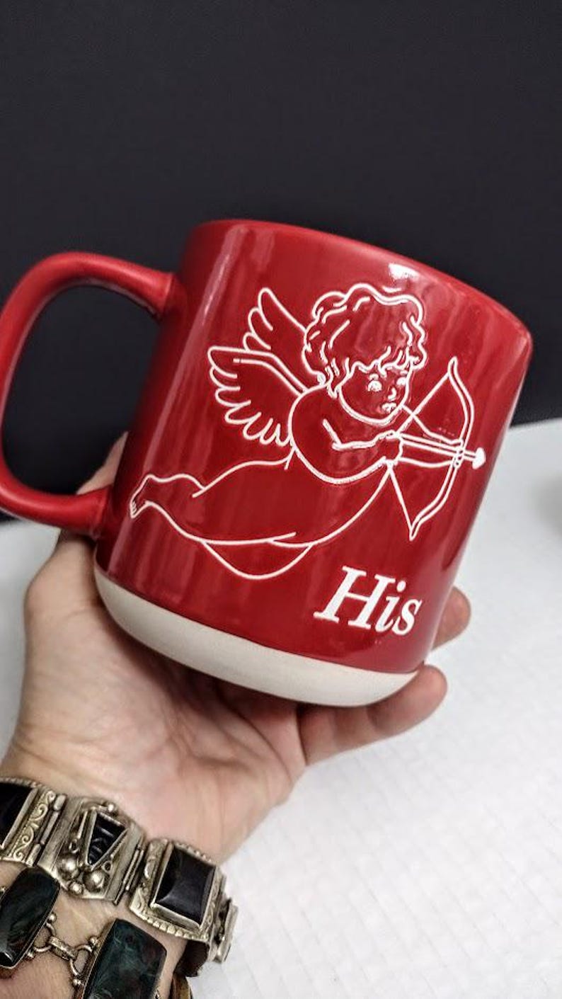 May include: Red ceramic mug with a white base and handle. The mug features a white illustration of Cupid with a bow and arrow, and the word "His" in white. The mug is held in a hand.