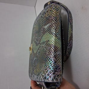 May include: A shoulder bag featuring a multi-coloured snakeskin pattern with iridescent green, blue, and brown tones. The bag has a structured flap, a gold-toned metal turn-lock closure, and a matching shoulder strap with a textured scale finish.