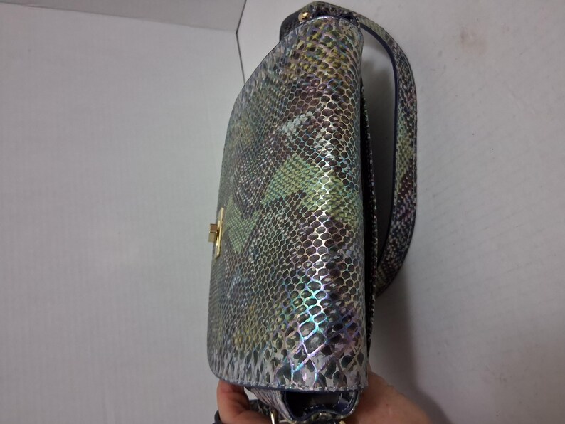 May include: A shoulder bag featuring a multi-coloured snakeskin pattern with iridescent green, blue, and brown tones. The bag has a structured flap, a gold-toned metal turn-lock closure, and a matching shoulder strap with a textured scale finish.