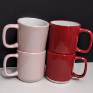 May include: Four ceramic mugs in a stack. Two are a light pink color, and two are a deep red. Each mug has a white interior and a large, curved handle. The mugs are stacked in pairs, with one pink and one red mug on top of each other.
