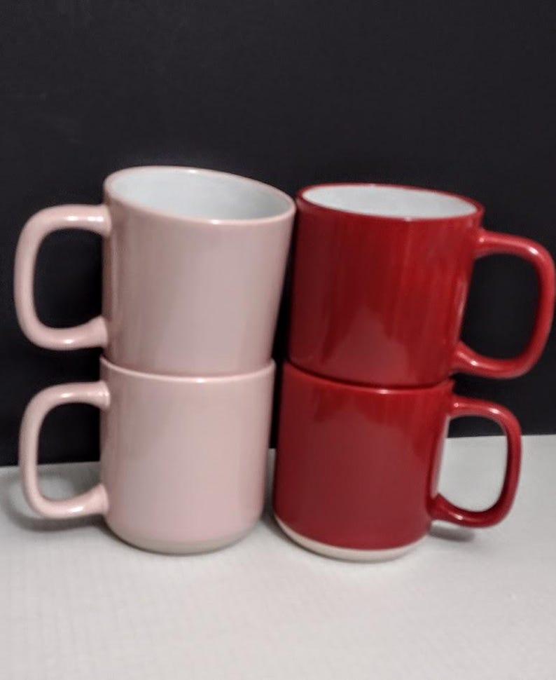 May include: Four ceramic mugs in a stack. Two are a light pink color, and two are a deep red. Each mug has a white interior and a large, curved handle. The mugs are stacked in pairs, with one pink and one red mug on top of each other.