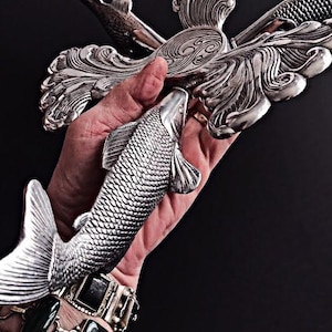 May include: A silver-toned metal object featuring a central circular medallion with a swirling pattern, surrounded by three sculpted fish figures with detailed scales and fins, arranged in a radial design with ornate, flowing leaf-like elements.