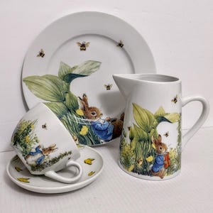 May include: A white ceramic tea set featuring a whimsical design with a rabbit in a blue jacket, green foliage, yellow flowers, and bees. The set includes a plate, a pitcher, a teacup, and a saucer.