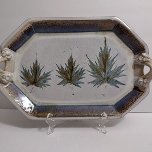 May include: An octagonal ceramic serving tray with two side handles. The tray features a speckled off-white base with three dark green and brown maple leaf imprints in the center. The rim is accented with a dark blue and brown glazed border.