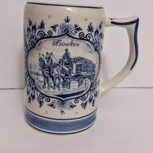 May include: A white ceramic beer stein with blue floral designs and a blue rim. The stein features a blue and white illustration of a horse-drawn carriage with the word "Heineken" above the image.