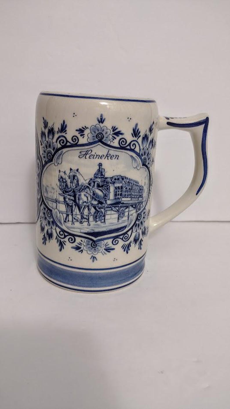 May include: A white ceramic beer stein with blue floral designs and a blue rim. The stein features a blue and white illustration of a horse-drawn carriage with the word "Heineken" above the image.