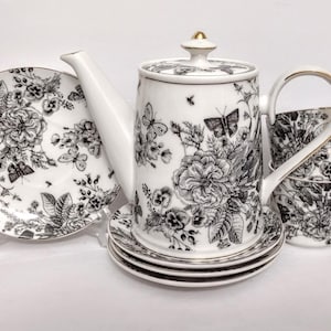 May include: A white porcelain tea set with a black floral and butterfly design. The set includes a teapot, two teacups, saucers, and plates. The teapot has a gold-colored handle and lid detail. The plates are supported by a clear plastic stand.