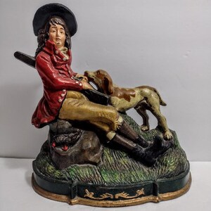 May include: A cast iron doorstop featuring a figure in a red coat and black hat sitting on a rock with a hunting dog. The figure holds a long black rifle across their lap. The base is painted green with gold trim and textured grass details.