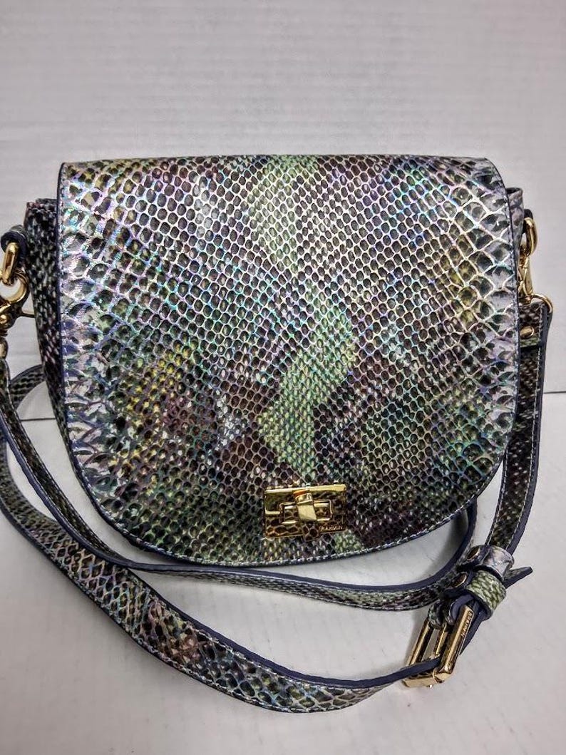 May include: A saddle-shaped crossbody bag featuring an iridescent snakeskin pattern in shades of green, purple, and silver. The bag includes a gold-tone rectangular twist-lock closure on the front flap and an adjustable shoulder strap with gold hardware.