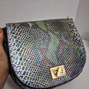 May include: A saddle-shaped shoulder bag featuring a multi-coloured iridescent snakeskin-embossed pattern in shades of green, blue, and black. The bag includes a gold-tone metal turn-lock closure with the brand name Brahmin engraved on the hardware.