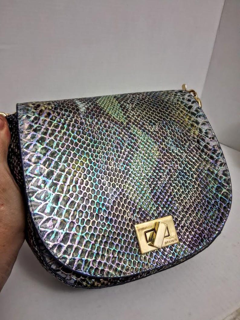 May include: A saddle-shaped shoulder bag featuring a multi-coloured iridescent snakeskin-embossed pattern in shades of green, blue, and black. The bag includes a gold-tone metal turn-lock closure with the brand name Brahmin engraved on the hardware.