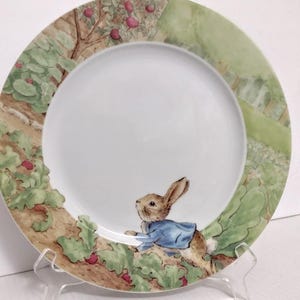 May include: A white ceramic plate with a colorful illustration of Peter Rabbit. The plate's border features a detailed scene of a garden with trees, apples, and foliage. Peter Rabbit is depicted wearing a blue jacket.