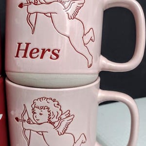 May include: Two pink ceramic mugs stacked on top of each other. Each mug features a red outline of Cupid with a bow and arrow, and the word "Hers" in red. The mugs have a curved handle and a light gray base.
