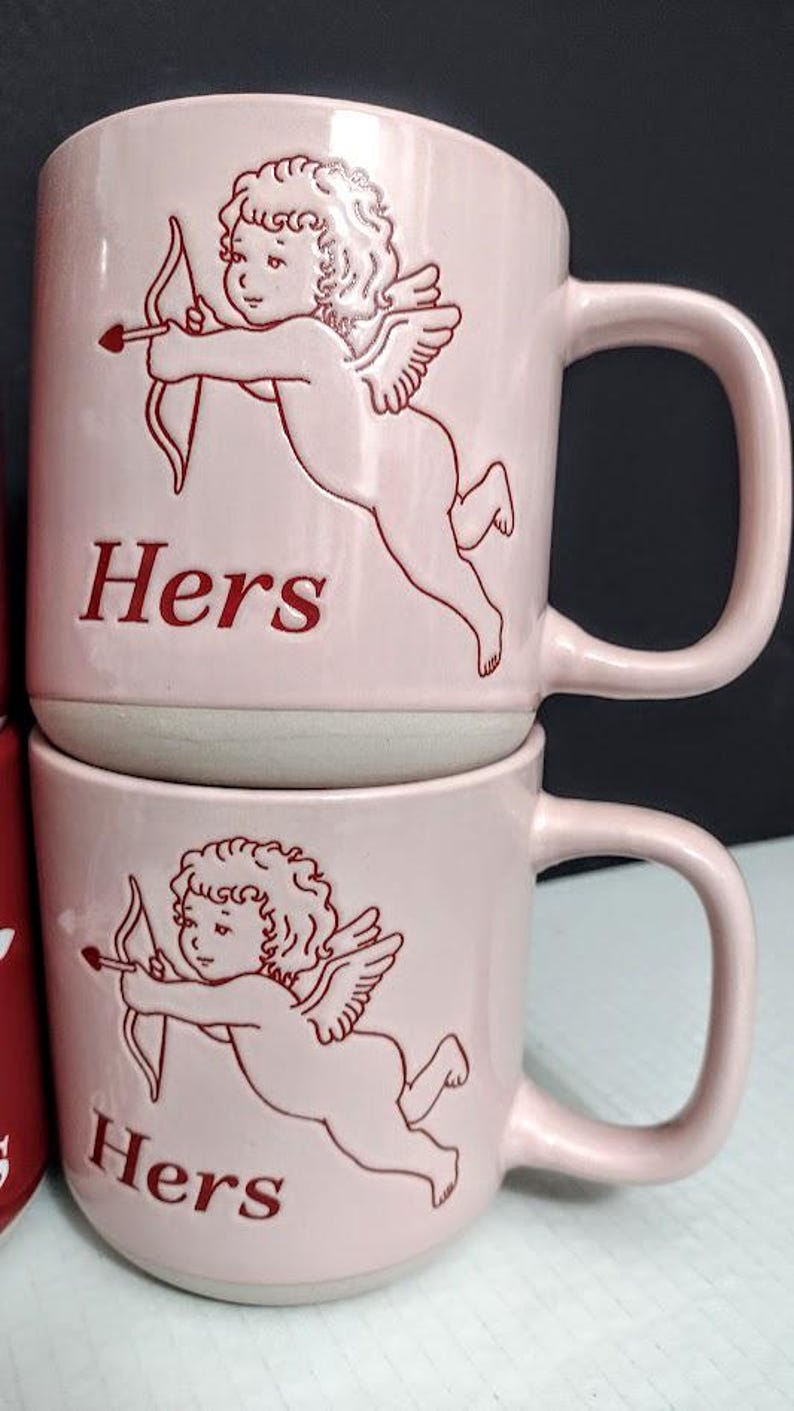 May include: Two pink ceramic mugs stacked on top of each other. Each mug features a red outline of Cupid with a bow and arrow, and the word "Hers" in red. The mugs have a curved handle and a light gray base.