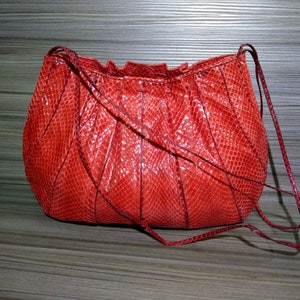 May include: A bright red handbag featuring a textured snakeskin pattern with a glossy finish. The bag has a rounded, pleated body design with a gathered top opening and thin, matching red shoulder straps attached to the sides of the structure.