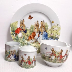 May include: A white ceramic dish set featuring illustrations of rabbits in a garden setting. The set includes a large plate, a small bowl, a creamer, and a lidded jar. The rabbits are depicted in various outfits, with butterflies and flowers.