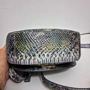 May include: A crossbody bag featuring a textured snakeskin pattern in iridescent shades of green, blue, and silver. The bag has a rounded, structured base with a metallic gold-tone clasp closure and a matching adjustable shoulder strap.
