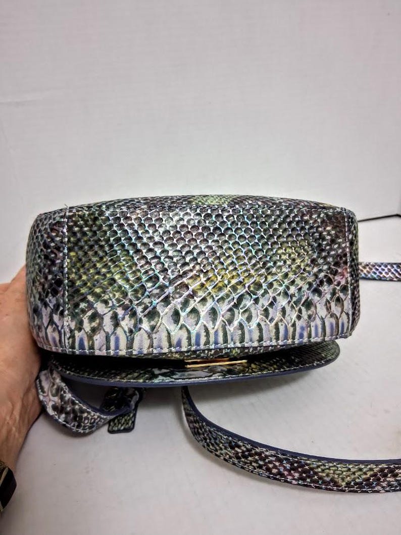May include: A crossbody bag featuring a textured snakeskin pattern in iridescent shades of green, blue, and silver. The bag has a rounded, structured base with a metallic gold-tone clasp closure and a matching adjustable shoulder strap.