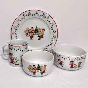 May include: A four-piece ceramic dish set featuring a festive holiday design. The set includes a large plate, two bowls, and a mug. Each piece is white with a red trim and decorated with a cartoon group of children and a Christmas tree.